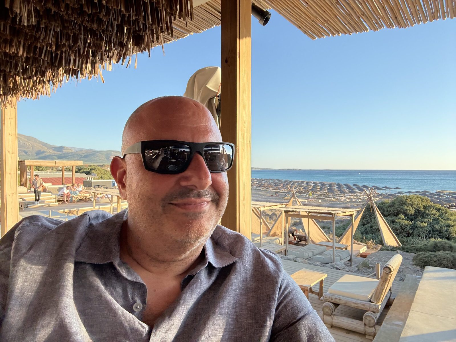 Emmanuel Tsouris in sunglasses and a linen shirt, sitting under a thatched beach shade with sunbeds and the sea behind him