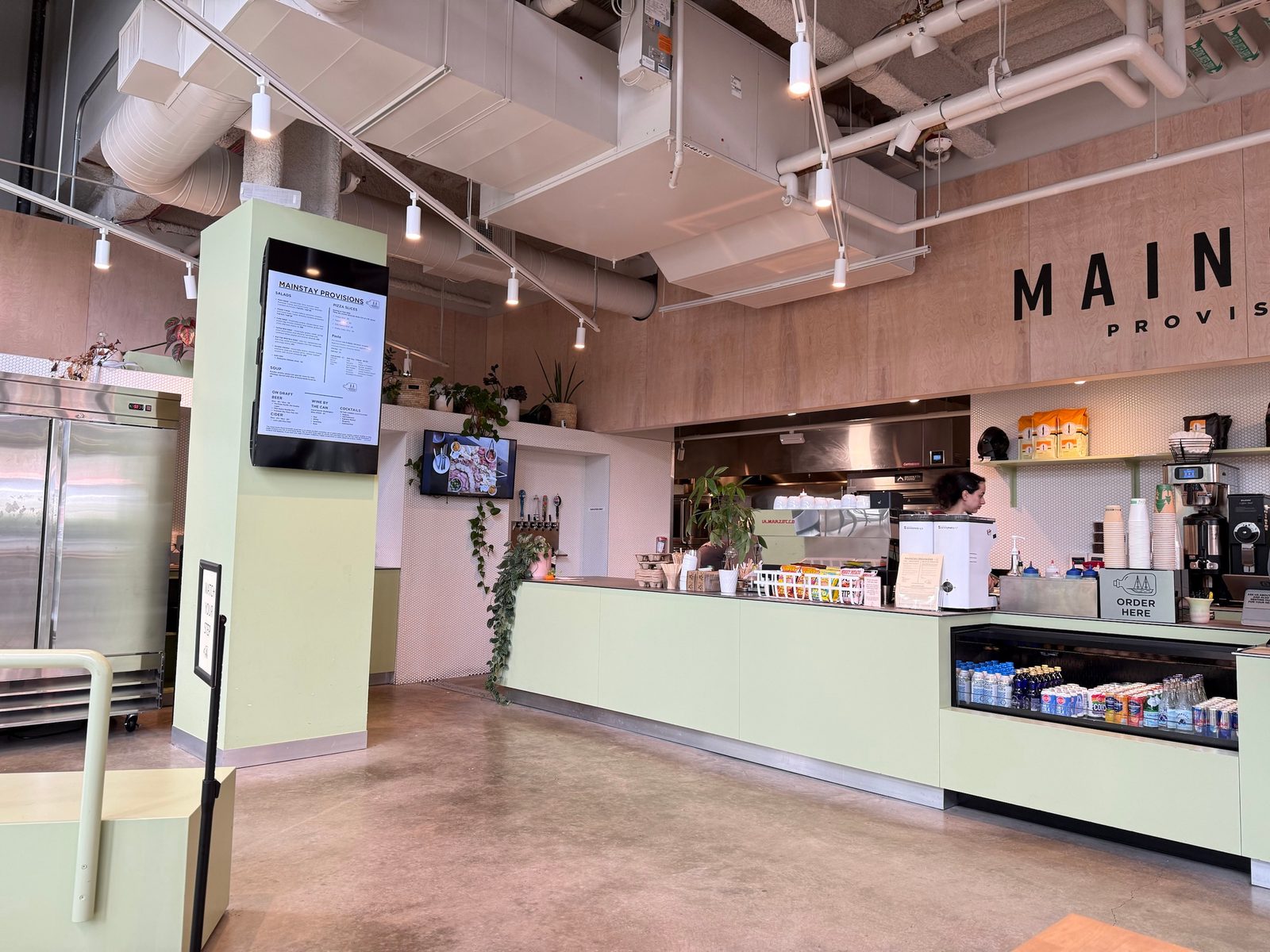 The Mainstay Provisions counter: a pale green service counter with an ORDER HERE sign, an espresso machine and a drinks case, under exposed ductwork, with the menu on a screen mounted to a green pillar