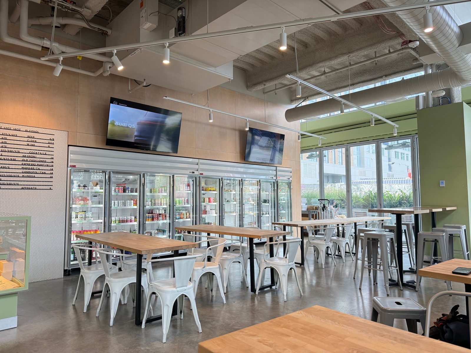 The Mainstay dining room: butcher-block tables with white metal chairs, a wall of glass-door drink coolers, two televisions, and a row of tall stools by folding glass windows