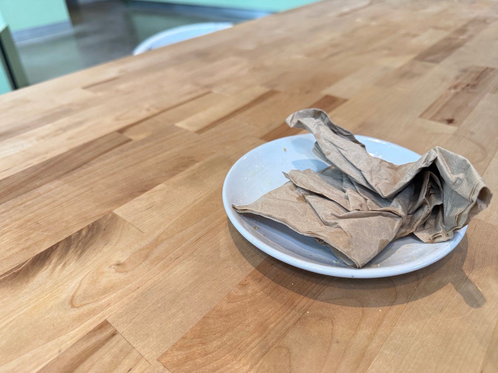 An empty white plate on a butcher-block table holding a crumpled pile of brown paper napkins and a few crumbs