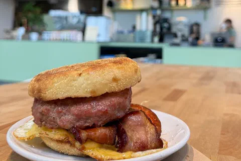 A breakfast sandwich on a small white plate on a butcher-block table: a toasted English muffin over a thick sausage patty, folded bacon, a fried egg and melted American cheese, with the MAINSTAY sign and the counter blurred in the background