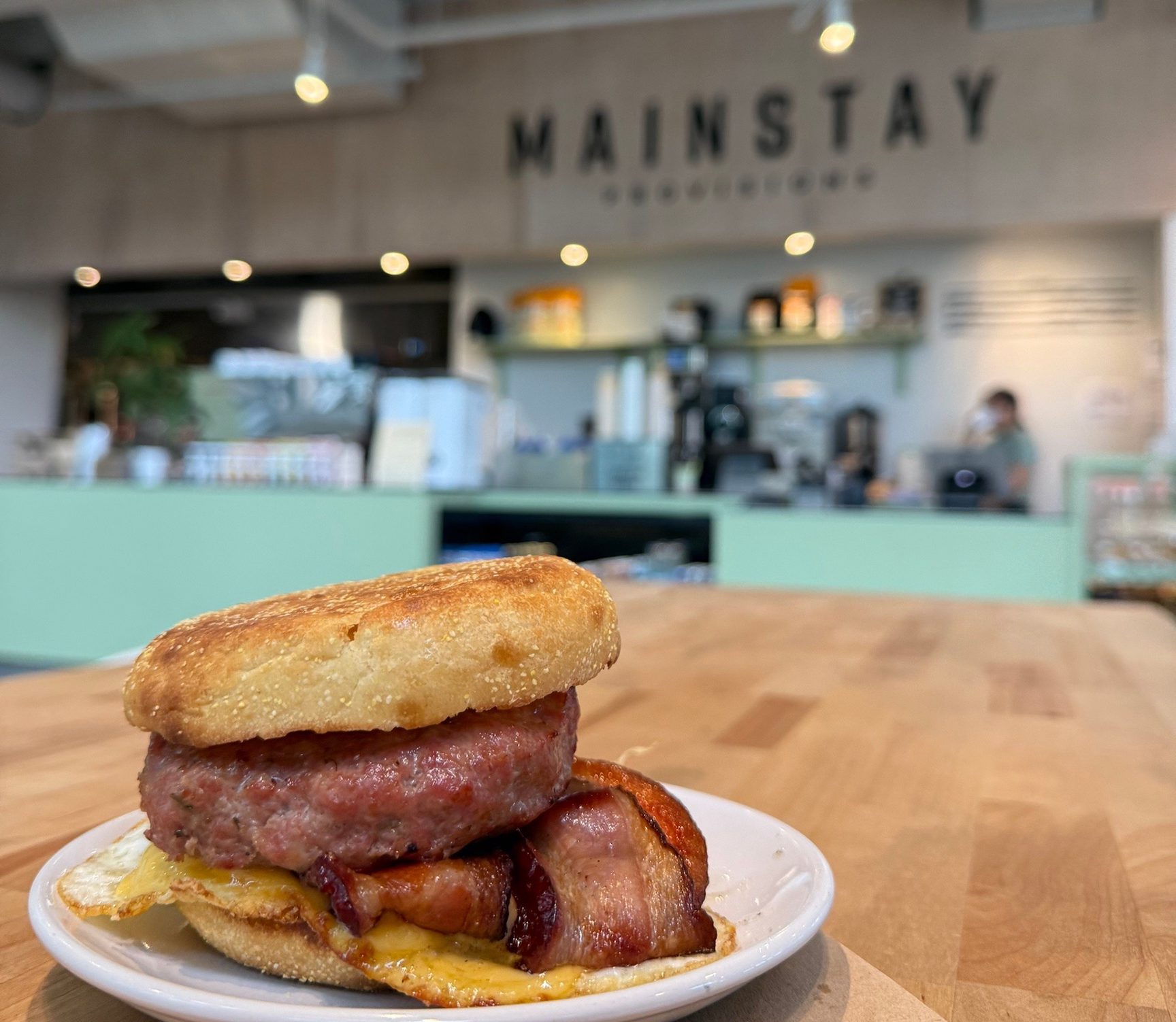A breakfast sandwich on a small white plate on a butcher-block table: a toasted English muffin over a thick sausage patty, folded bacon, a fried egg and melted American cheese, with the MAINSTAY sign and the counter blurred in the background