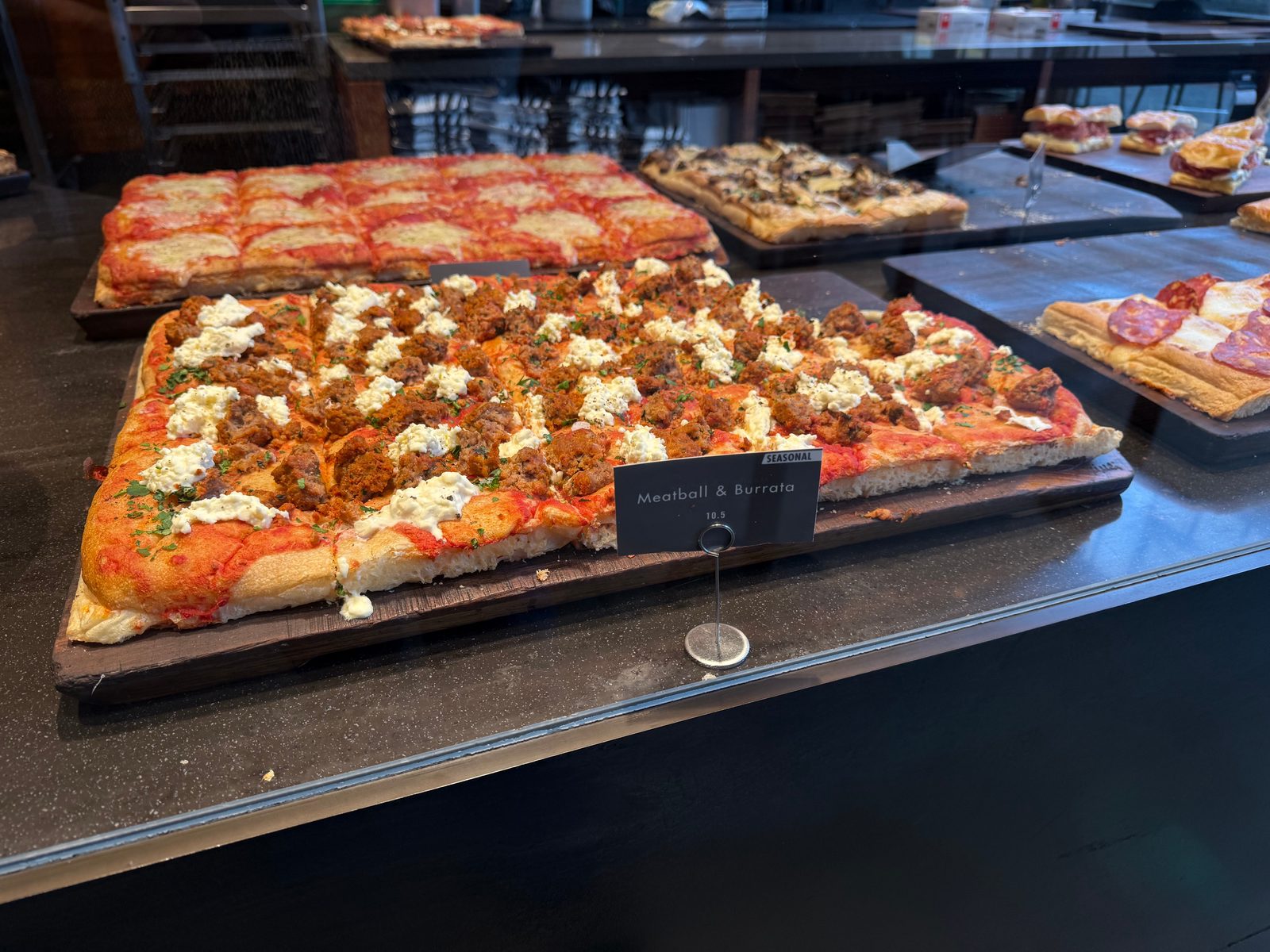 Rectangular pizzas behind the bakery glass, with a meatball and burrata pizza in the foreground.