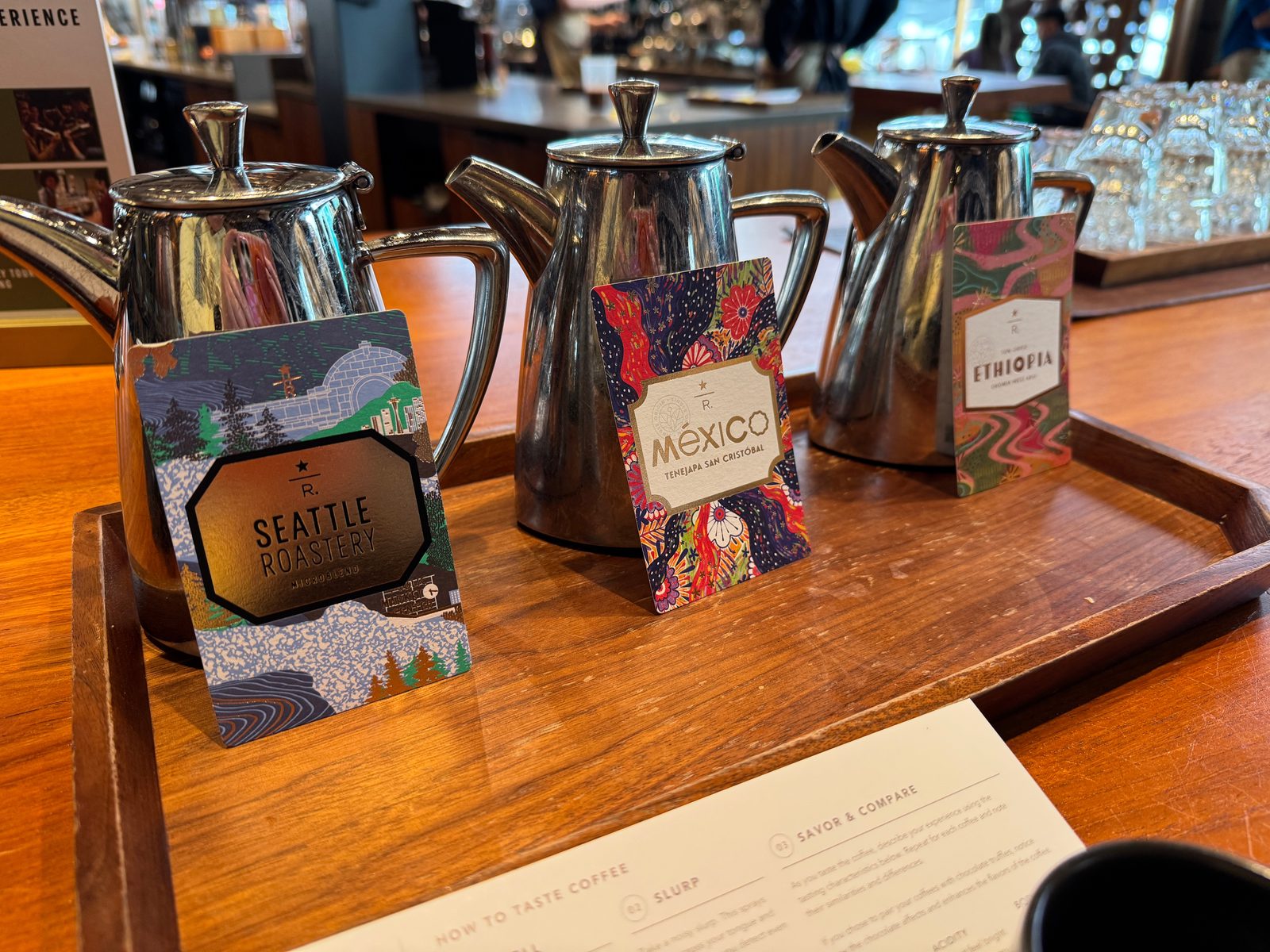 Three metal coffee pots on a wooden tray with cards marked Seattle Roastery, México and Ethiopia, beside a coffee-tasting guide.