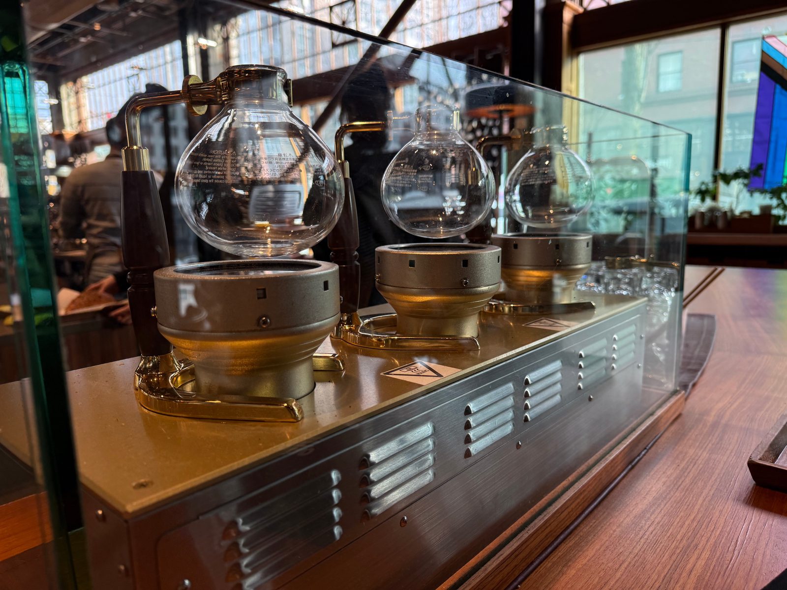 Three glass siphon brewers on metal heating stands behind a glass screen at the coffee counter.