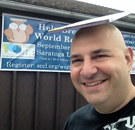 Close selfie of a smiling man with a hardback book balanced flat on his head, in front of a library banner reading “Help Break a World Record, September 2014, Saratoga Library” and “Register: sccl.org/worldrecord”