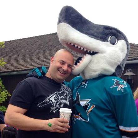 A man in a black T-shirt holding a paper coffee cup, leaning cheek to cheek with S.J. Sharkie, the San Jose Sharks mascot, who wears a teal Sharks jersey and a large grey-and-white shark head