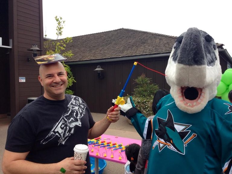 The same man with a hardback book balanced flat on his head, still holding the coffee, beside S.J. Sharkie holding a toy fishing rod over a children’s magnetic fishing game on a pink table