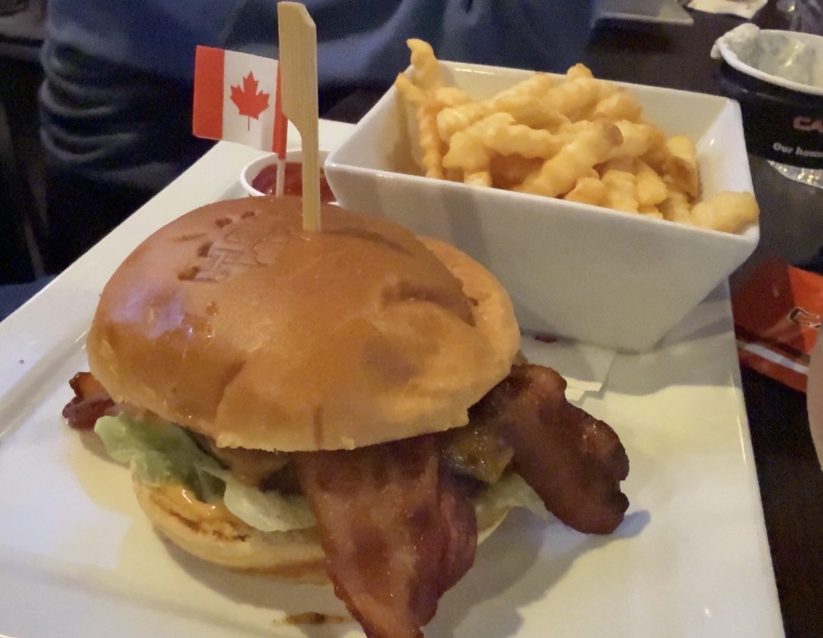 A bacon cheeseburger on a branded bun with crinkle-cut fries in a white bowl, a small Canadian flag pick standing in the burger