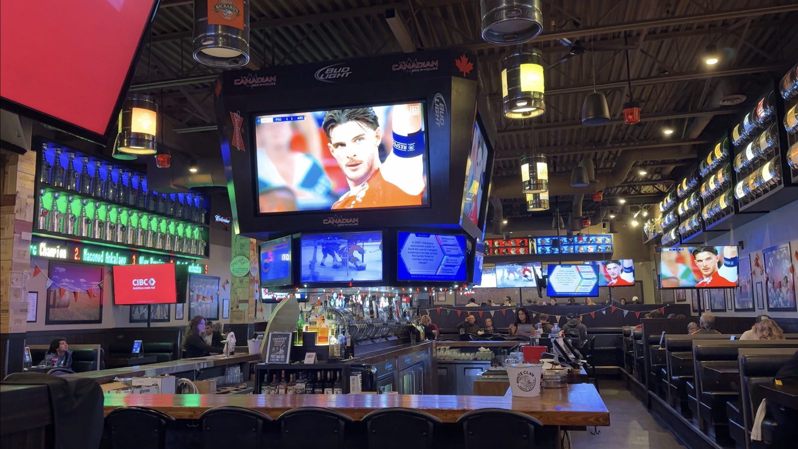 A large sports pub interior with a central bar, many screens showing hockey and soccer, kegs used as pendant light fixtures, and bottle displays lit in blue and green along the walls