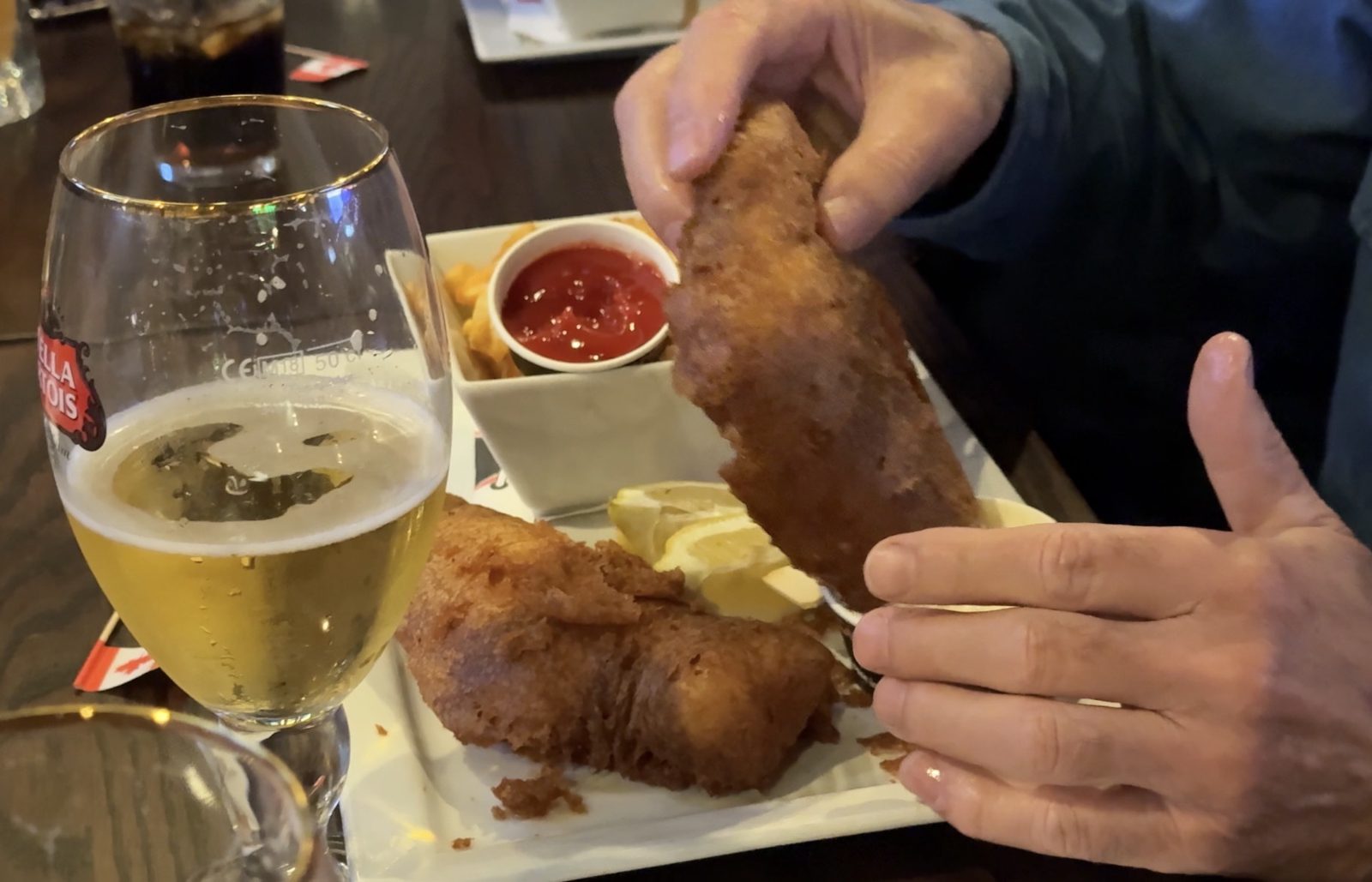 A hand lifting a large battered fish fillet from a plate with fries, ketchup and a lemon wedge, beside a glass of lager