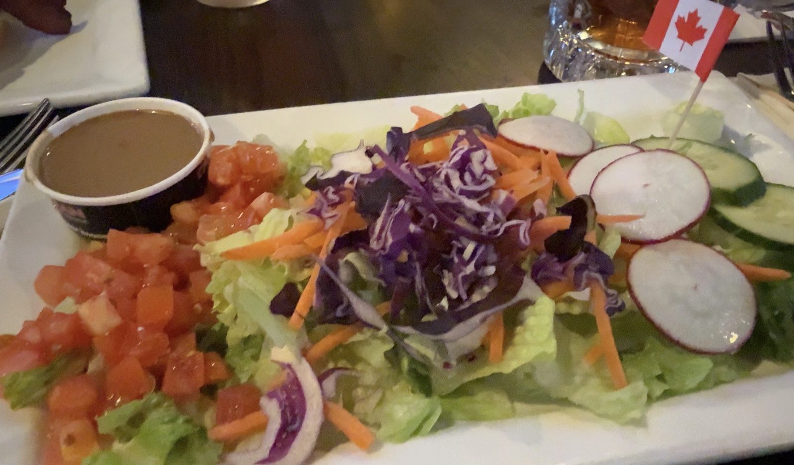 A side salad on a rectangular white plate — lettuce, diced tomato, cucumber, radish, shredded carrot and red cabbage, with balsamic dressing in a ramekin and a Canadian flag pick