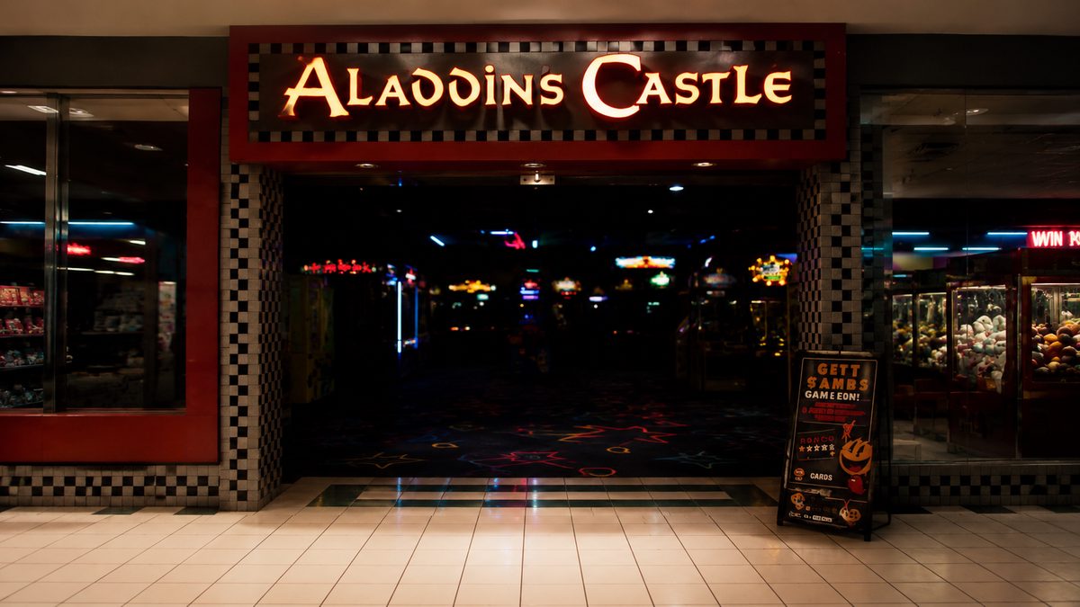 The entrance to an Aladdin's Castle arcade in a dim mall, with an illuminated red and yellow sign above a checkered archway and the neon glow of cabinets behind it.