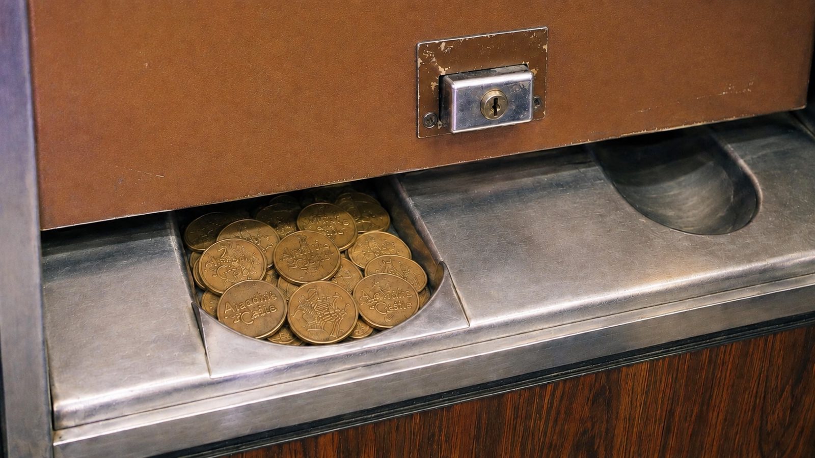 Brass Aladdin’s Castle tokens collected inside a recessed payout cup beneath the brown panel of a change machine with a stainless-steel ledge and woodgrain base.