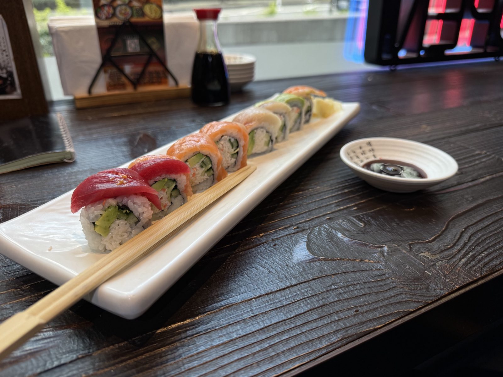 A rainbow roll on a long white plate, tuna and salmon over the top, chopsticks resting across the plate and a dish of soy sauce beside it