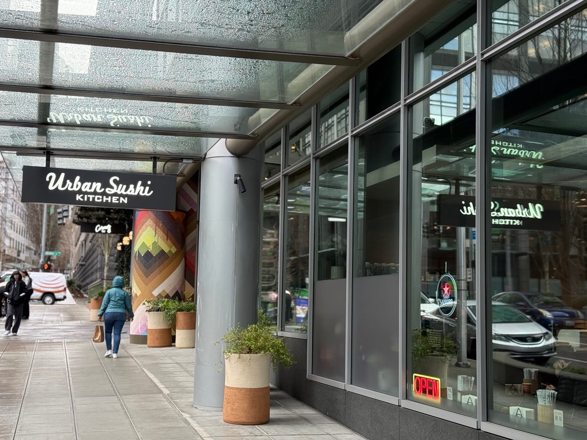 The Urban Sushi Kitchen storefront under a glass awning on a wet sidewalk, floor-to-ceiling windows with a Sapporo sign and a red OPEN sign inside